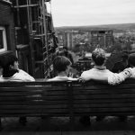 Four friends sitting together on a bench overlooking a city skyline, seen from behind — a quiet moment of human connection